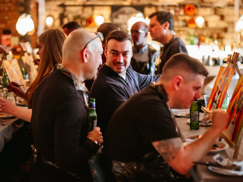 Three men at a paint and sip class with people in the background